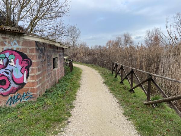 Chemin de terre bordé d'une clôture sur la droite. Sur la gauche, un bâtiment en briques avec des graffitis.